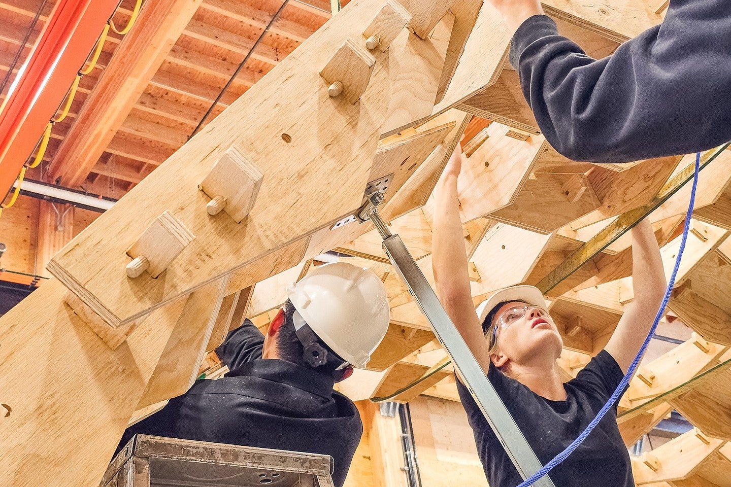 Students wear hard hats as they assemple wooden architectural structure