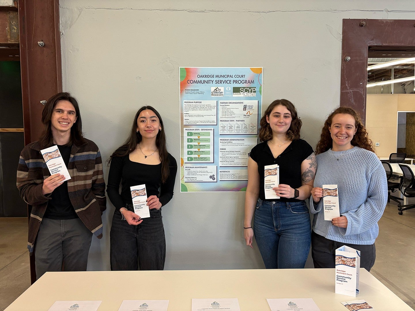 Four students smile in front of their class presentation poster