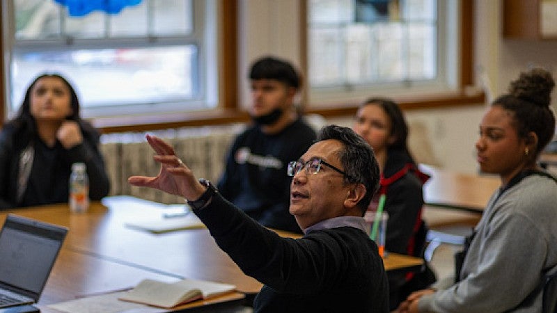 Professor DeVigal sits at table with students and teaches a lesson