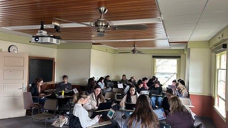 Students sit at desks and gather in teams while they work on laptops