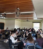 Students sit at desks and gather in teams while they work on laptops